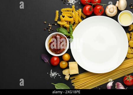 Assiette entourée de différents types de pâtes, produits et épices sur fond noir, plat. Espace pour le texte Banque D'Images
