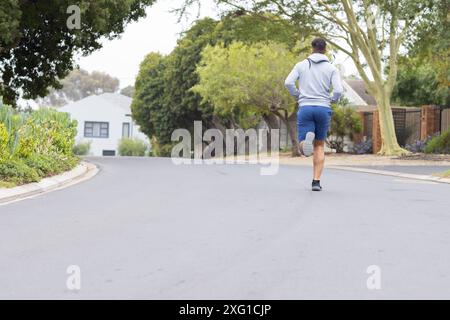 Jogging sur la rue de banlieue, homme en sweat à capuche et short s'exerçant à l'extérieur, espace de copie Banque D'Images