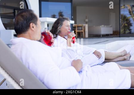 Détente au bord de la piscine, couple senior dans des peignoirs buvant des boissons rouges en vacances Banque D'Images