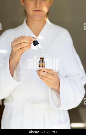 Tenant compte-gouttes et bouteille d'huile essentielle, femme en peignoir préparant la routine de soins de la peau Banque D'Images