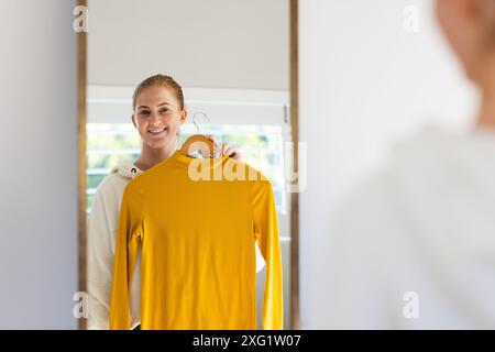 Femme souriante tenant un pull jaune devant le miroir, décidant de la tenue Banque D'Images