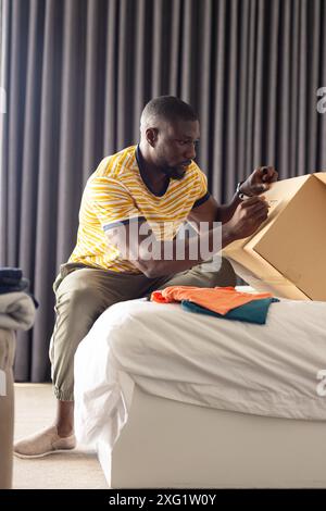 Ouverture de la boîte en carton, homme assis sur le lit dans la chambre, déballage des vêtements Banque D'Images