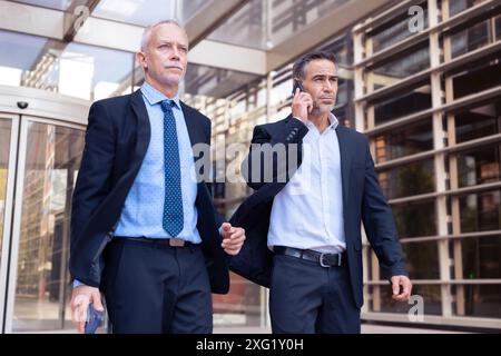 Deux hommes d'affaires heureux marchant à l'extérieur de l'immeuble de bureaux avec smartphone à la main Banque D'Images