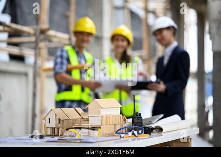 Un modèle architectural, des plans et des outils sur la table avec des ouvriers de la construction debout dans un fond flou Banque D'Images