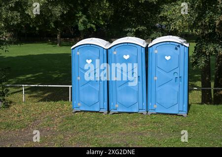 Trois toilettes mobiles bleues dans le parc lors d'un événement de festival en plein air sur la prairie verte sous les arbres dans un parc de campagne publique, espace copie, sélectionné Banque D'Images