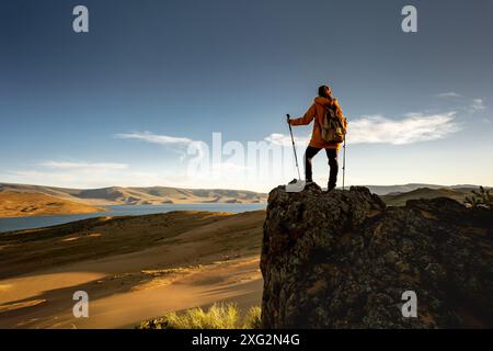 Femme randonneur avec bâtons de randonnée et sac à dos est debout sur le grand rocher contre le désert de coucher de soleil, ciel bleu et lac Banque D'Images