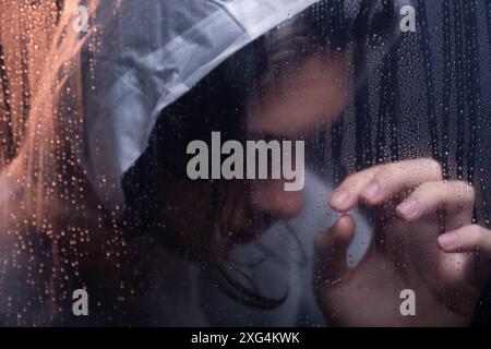 Portrait en gros plan d'une jeune femme portant un imperméable en plastique transparent derrière du verre. Prise de vue en studio. Isolé sur fond sombre. Banque D'Images