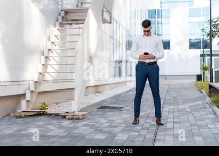 Un homme portant une chemise blanche et un pantalon bleu se tient debout et regarde son téléphone portable, se concentrant intensément sur l'écran. Il semble engagé et absorbé. Banque D'Images