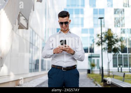 Un homme portant une chemise blanche et un pantalon bleu se tient debout et regarde attentivement un téléphone portable entre ses mains. Banque D'Images