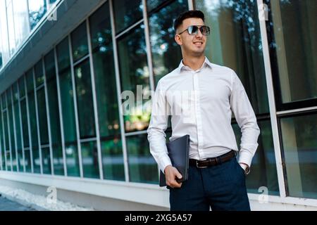 Un homme portant des lunettes de soleil sombres se tient confiant devant un bâtiment moderne. Il semble observer son environnement avec un comportement composé. Banque D'Images