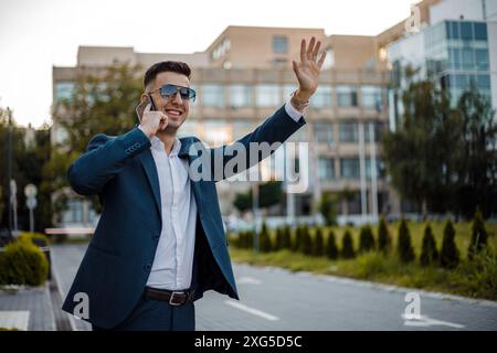 Un jeune homme en costume bleu et lunettes de soleil se tient dans une rue de la ville, parlant sur un téléphone portable tout en levant le bras pour héler un taxi. Banque D'Images