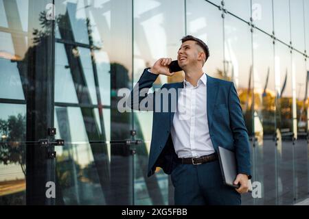 Un homme d'affaires en costume bleu et chemise blanche se tient devant un immeuble de bureaux en verre moderne tout en parlant sur son smartphone. Banque D'Images