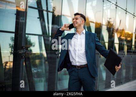 Un homme portant un costume et une chemise blanche se tient devant un bâtiment moderne en verre. Il tient un ordinateur portable sous un bras et parle au téléphone. Banque D'Images