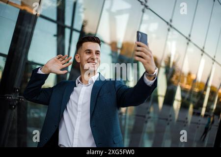 Un jeune homme en costume bleu et chemise blanche se tient devant un bâtiment en verre et prend un selfie avec son téléphone. Il sourit et agite devant la caméra. Banque D'Images