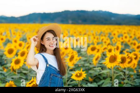femme asiatique avec salopettes jeans et chapeau de rotin souriant sur le champ de tournesol dans le concept de belle nature Banque D'Images