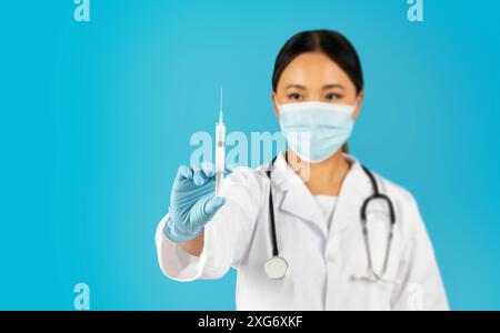 Femme médecin portant le masque facial tient la seringue sur fond bleu Banque D'Images