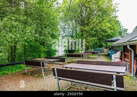 Terrasse arrière rustique d'un restaurant de campagne, tables et bancs en bois, chaises en plastique empilées à côté du mur, arbres feuillus avec feuillages verts en backgro Banque D'Images