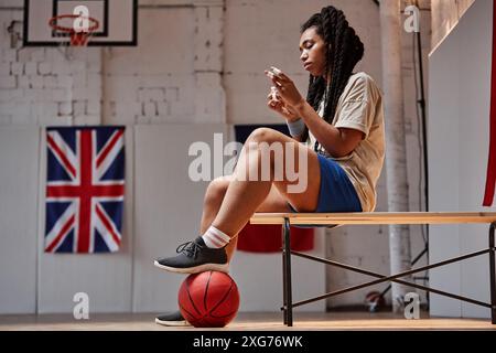 Portrait de vue de côté de la jeune femme noire en tant que joueuse de basket-ball se préparant pour le match assis sur le banc dans la cour intérieure avec des drapeaux dans l'espace de copie d'arrière-plan Banque D'Images