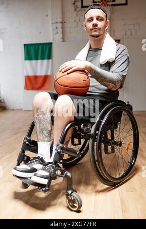 Portrait vertical de l'homme adulte tatoué avec un handicap en fauteuil roulant dans un terrain de basket-ball intérieur et en regardant la caméra avec une lumière dramatique Banque D'Images