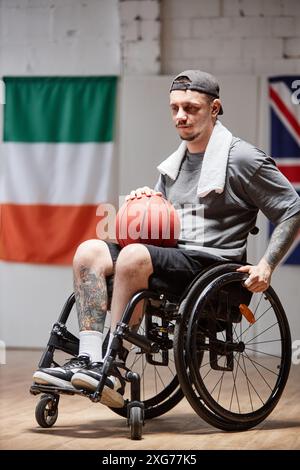 Portrait vertical pleine longueur de joueur de basket tatoué avec handicap en fauteuil roulant dans un court intérieur avec éclairage aérien Banque D'Images