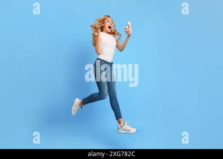 Femme émotionnelle sautant tout en regardant smartphone sur fond bleu Banque D'Images