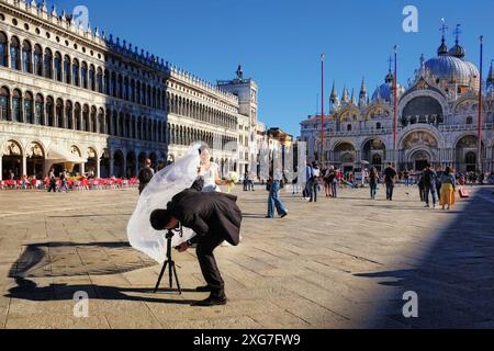 Une mariée et un marié travaillant ensemble sur la place Saint-Marc à Venise, le goom place la caméra tenant le voile blanc des mariées, le travail d'équipe fait des photos. Banque D'Images