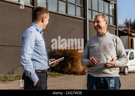 Deux hommes se tiennent debout à l'extérieur d'un bâtiment industriel, engagés dans la conversation. Banque D'Images