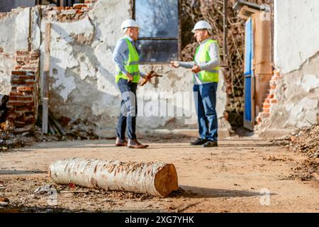 Deux hommes portant des casques de sécurité et des gilets réfléchissants se tiennent devant un bâtiment partiellement démoli, discutant apparemment des plans pour le projet de construction. Banque D'Images