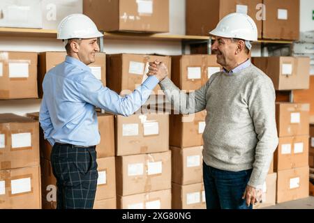 Deux hommes portant un casque blanc se tiennent devant un mur de boîtes empilées dans un entrepôt, serrant la main avec des sourires sur le visage. Banque D'Images