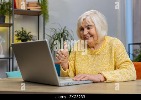 Femme senior faisant appel de conférence vidéo webcam avec des amis ou de la famille, montrer le pouce vers le haut, profiter d'une conversation agréable à la maison appartement à l'intérieur. Grand-mère âgée mature est assise sur le canapé à la table de bureau Banque D'Images