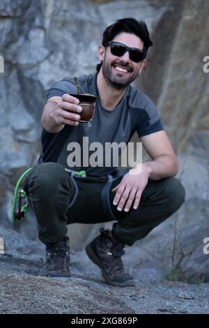 Portrait vertical d'un homme souriant accroupissant, portant des lunettes de soleil et du matériel de randonnée, profite d'un thé traditionnel au cours d'une aventure en montagne. Banque D'Images