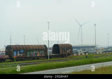 Gand, Belgique - 22 mai 2023 : deux wagons de train de marchandises portant des graffitis colorés sont stationnés sur les rails, avec une grande éolienne dans la distance brumeuse Banque D'Images