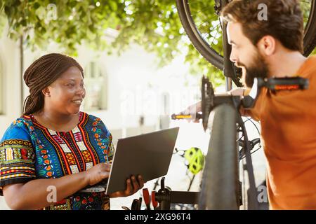 Jeune couple multiethnique actif et dynamique surfant sur Internet pour l'entretien du matériel de vélo. Amateurs de sport petit ami et petite amie entretien des pédales de vélo tout en utilisant un ordinateur portable pour le cyclisme d'été annuel. Banque D'Images