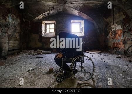 Triste homme assis dans un vieux fauteuil roulant dans un service hospitalier abandonné. Silhouette de personne dans un véhicule délabré pour malades handicapés dans un bâtiment ancien. Banque D'Images