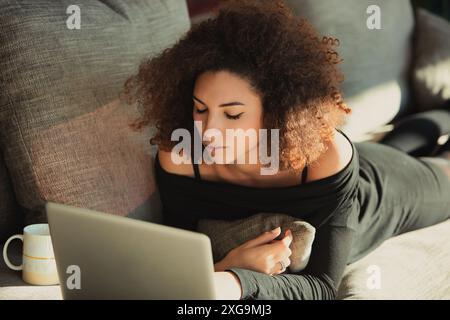 Jeune femme est allongée sur son canapé travaillant sur un ordinateur portable avec une tasse de café à proximité. Elle profite du confort de la maison tout en obtenant un peu de travail fait Banque D'Images
