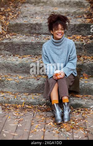 Heureuse jeune femme d'origine africaine dans des vêtements chauds assis dehors sur un escalier en pierre couvert de feuilles d'automne tombées, tenant tasse de chaud Banque D'Images