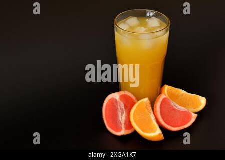 Un grand verre à facettes avec du jus fraîchement pressé avec l'ajout de glace sur un fond noir, à côté de tranches d'orange et de pamplemousse mûres. Gros plan. Banque D'Images