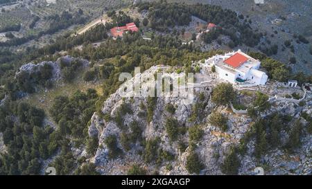 Drone shot, Un bâtiment sur une colline rocheuse, entouré par la nature boisée et d'autres bâtiments dans la région, église Panagia Kyra, au-dessus de la plage de Tsambika Banque D'Images