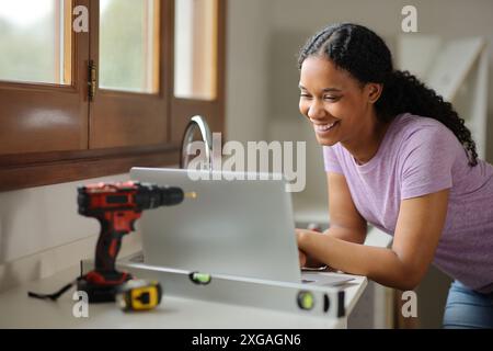 Femme noire heureuse vérifiant l'ordinateur portable reformant la maison dans la cuisine Banque D'Images