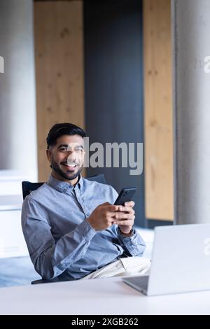Homme d'affaires souriant à l'aide de smartphone tout en étant assis au bureau avec ordinateur portable dans le bureau Banque D'Images