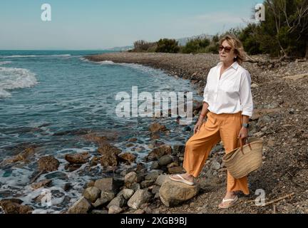 Une femme mature portant des lunettes de soleil et des vêtements décontractés se promène le long d'une plage rocheuse, profitant du paysage côtier serein par une journée ensoleillée. Banque D'Images