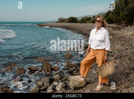 Une femme mature portant des lunettes de soleil et des vêtements décontractés se promène le long d'une plage rocheuse, profitant du paysage côtier serein par une journée ensoleillée. Banque D'Images