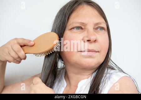peigne en bois, brosse dans la main féminine, femme mature heureuse peignant les longs cheveux bruns, enchevêtrements de cheveux tombés enroulés sur des épingles de brosse, concept perte de cheveux, santé, Banque D'Images