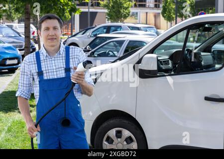 Un homme en uniforme tient la fiche de charge du véhicule électrique. Fourgon électrique en arrière-plan. Banque D'Images