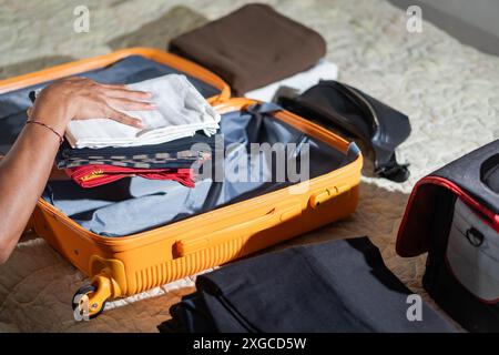 Une personne emballant des vêtements dans une valise orange ouverte sur un lit. Divers vêtements et une trousse de toilette noire sont visibles. Banque D'Images