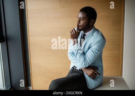 portrait de l'homme d'affaires afro-américain agréable à la mode habillé dans un manteau formel élégant assis au bureau avec un look pensif, thinkin Banque D'Images