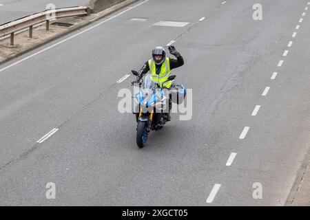 Motocycliste circulant le long d'une route principale avec une main levée ou agitée. Cycliste de moto portant un gilet haute visibilité. Banque D'Images