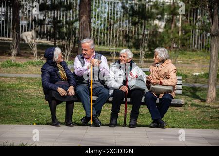 Plusieurs personnes retraitées sont assises sur un banc dans un parc public de la ville, profitant du temps ensoleillé, bavardant et se relaxant, entourées de verdure et d'arbres Banque D'Images