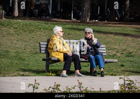 Plusieurs personnes retraitées sont assises sur un banc dans un parc public de la ville, profitant du temps ensoleillé, bavardant et se relaxant, entourées de verdure et d'arbres Banque D'Images