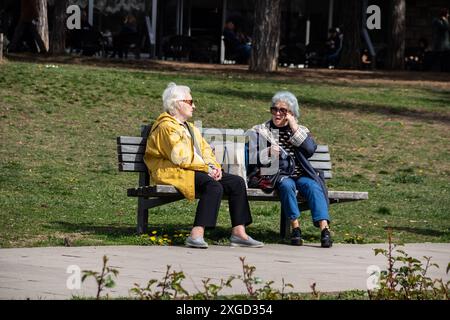 Plusieurs personnes retraitées sont assises sur un banc dans un parc public de la ville, profitant du temps ensoleillé, bavardant et se relaxant, entourées de verdure et d'arbres Banque D'Images